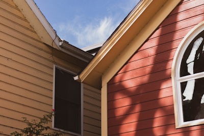 Two house exteriors with siding are visible, one beige and the other red. The red house features a large arched window. Soft shadows of nearby trees are cast on the siding. The sky is clear and blue.