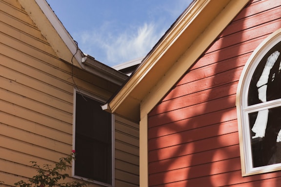 Two house exteriors with siding are visible, one beige and the other red. The red house features a large arched window. Soft shadows of nearby trees are cast on the siding. The sky is clear and blue.