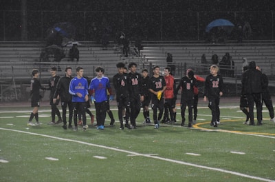A group of soccer players wearing black and blue uniforms walks on a wet field during a rainy night. The players seem to be finishing a game, and the stands in the background are mostly empty, with a few spectators holding umbrellas.