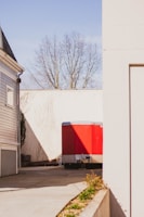 A red utility trailer is parked on a paved driveway between two buildings. The building on the left is a light gray house with white trim. On the right is a white building with a flat wall. In the background, there is a white fence and leafless tree branches are visible above the fence.