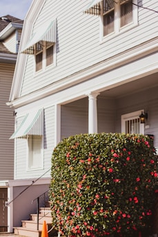 A well-maintained exterior of a house with light-colored siding and green-and-white striped awnings above the windows. A large, neatly trimmed shrub with numerous red flowers is positioned near the entrance, next to a small set of stairs leading to the front door. The bright sunlight casts soft shadows, suggesting a clear day.