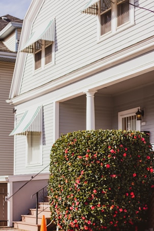 A well-maintained exterior of a house with light-colored siding and green-and-white striped awnings above the windows. A large, neatly trimmed shrub with numerous red flowers is positioned near the entrance, next to a small set of stairs leading to the front door. The bright sunlight casts soft shadows, suggesting a clear day.