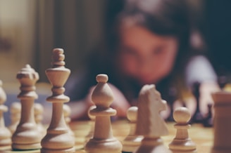 A close-up view of chess pieces on a board, with the white pieces prominently displayed and in focus. In the background, a person appears blurred, suggesting intense concentration or contemplation.