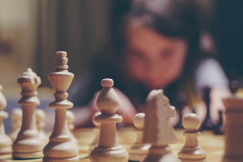 A close-up view of chess pieces on a board, with the white pieces prominently displayed and in focus. In the background, a person appears blurred, suggesting intense concentration or contemplation.