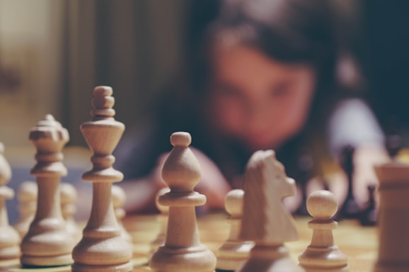 A close-up view of chess pieces on a board, with the white pieces prominently displayed and in focus. In the background, a person appears blurred, suggesting intense concentration or contemplation.