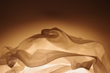 Softly illuminated translucent fabric folds create a warm and gentle play of light and shadow against a smooth background. The fabric appears to be lightly layered, forming gentle peaks and valleys.