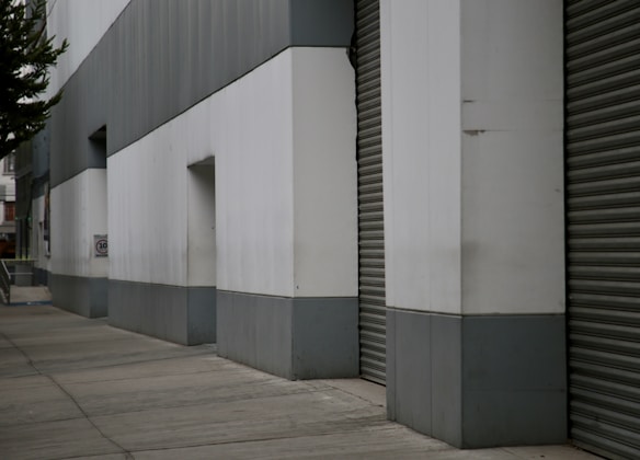 A street with a large gray and white industrial building featuring flat, metal panels and large garage-style doors. The sidewalk is concrete, with some greenery visible on the left side. The architecture appears utilitarian with clean lines and minimalistic design.