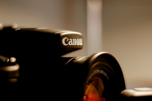 A close-up view of a black Canon camera with focus on the brand logo and part of the lens. The background is blurred, giving an emphasis on the camera itself.