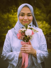 A person is wearing a light blue hijab and a matching outfit with decorative buttons, holding a bouquet of flowers with light pink and white blooms. The bouquet is tied with a pink ribbon, and the person is smiling gently against a background of green foliage.