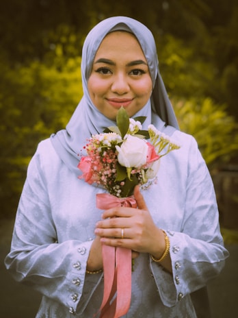A person is wearing a light blue hijab and a matching outfit with decorative buttons, holding a bouquet of flowers with light pink and white blooms. The bouquet is tied with a pink ribbon, and the person is smiling gently against a background of green foliage.