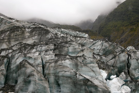 person in blue jacket sitting on rock mountain during daytime in Fox Glacier New Zealand