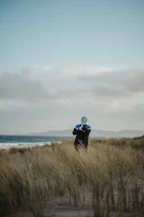 A person stands on a sandy beach surrounded by tall grass, holding a paddle vertically. The ocean is visible to the left, with waves gently crashing on the shore. The sky is a soft blue with scattered clouds, creating a serene atmosphere.