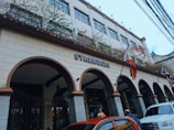 A building with Spanish-style architecture featuring arched entryways and a tiled roof, adorned with Starbucks signage. Flags are displayed at the front, including the Philippine flag. Vines with flowers embellish the upper part of the structure. Several cars are parked in front of the building, and people are visible near the entrance.