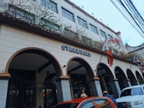 A building with Spanish-style architecture featuring arched entryways and a tiled roof, adorned with Starbucks signage. Flags are displayed at the front, including the Philippine flag. Vines with flowers embellish the upper part of the structure. Several cars are parked in front of the building, and people are visible near the entrance.