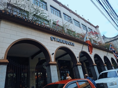 A building with Spanish-style architecture featuring arched entryways and a tiled roof, adorned with Starbucks signage. Flags are displayed at the front, including the Philippine flag. Vines with flowers embellish the upper part of the structure. Several cars are parked in front of the building, and people are visible near the entrance.