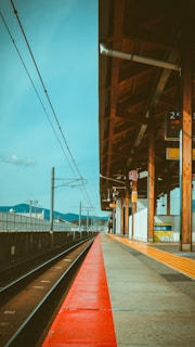A train station platform extending into the distance, with wooden beams supporting a slanted roof. The tracks run parallel to the platform, and the area is well-lit by natural daylight. There are signs and safety markings visible, with mountains in the background under a clear blue sky.