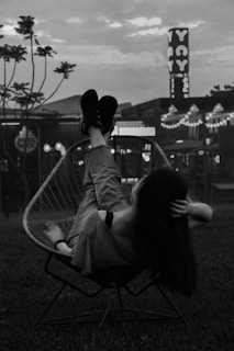 A person is reclining in a wire chair with their legs elevated, set against an urban backdrop featuring buildings and neon signs. The scene occurs during nighttime or dusk, lending a moody atmosphere with softly illuminated lights in the background. The silhouetted figure adds a sense of relaxation and calm.