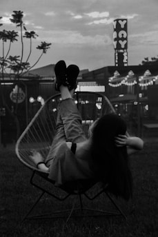 A person is reclining in a wire chair with their legs elevated, set against an urban backdrop featuring buildings and neon signs. The scene occurs during nighttime or dusk, lending a moody atmosphere with softly illuminated lights in the background. The silhouetted figure adds a sense of relaxation and calm.