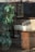 A bathroom setting is visible, featuring a white ceramic basin on a wooden cabinet. There is a large, leafy green plant beside the cabinet. Reflections on the glass suggest this is a window display, with outdoor elements visible, such as trees and a pavement.