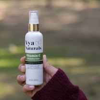 A hand is holding a bottle of Vya Naturals Vitamin C Moisturizer with a gold cap. The background is blurred, but appears to be outdoor and green.