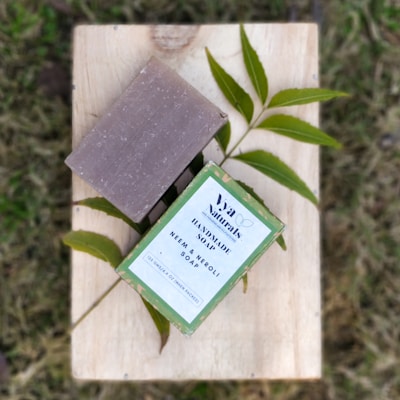 A bar of handmade soap and its packaging are placed on a wooden surface with green leaves underneath. The packaging is labeled with 'Va Naturals Handmade Soap Neem & Neroli', and the natural elements surrounding the soap enhance its eco-friendly appeal.