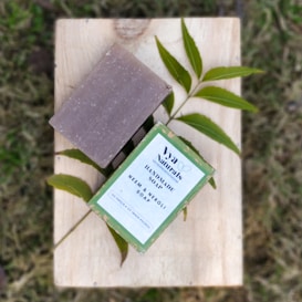A bar of handmade soap and its packaging are placed on a wooden surface with green leaves underneath. The packaging is labeled with 'Va Naturals Handmade Soap Neem & Neroli', and the natural elements surrounding the soap enhance its eco-friendly appeal.
