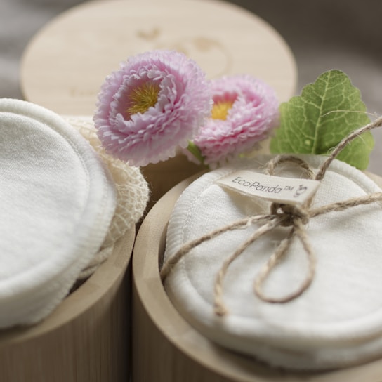 A close-up view of two round wooden containers, one with a lid and the other holding soft, white reusable fabric pads tied with a string. Adjacent to the pads, light pink flowers and a green leaf provide a touch of natural aesthetics.