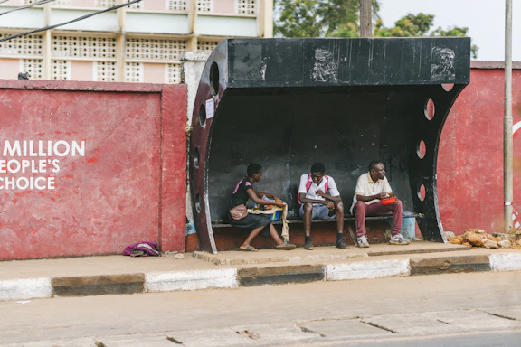 A mother with two children waiting at a bus stop in front of modest housing, capturing the daily reality of relying on public transit.