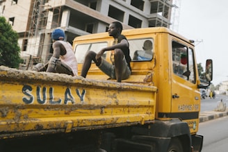 A yellow construction truck is on the road, with two young men sitting casually in the back. The truck is slightly weathered and has the word 'SULAY' painted on the side. A large building under construction is visible in the background, along with some scaffolding.
