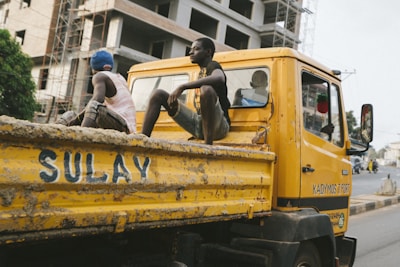 A yellow construction truck is on the road, with two young men sitting casually in the back. The truck is slightly weathered and has the word 'SULAY' painted on the side. A large building under construction is visible in the background, along with some scaffolding.
