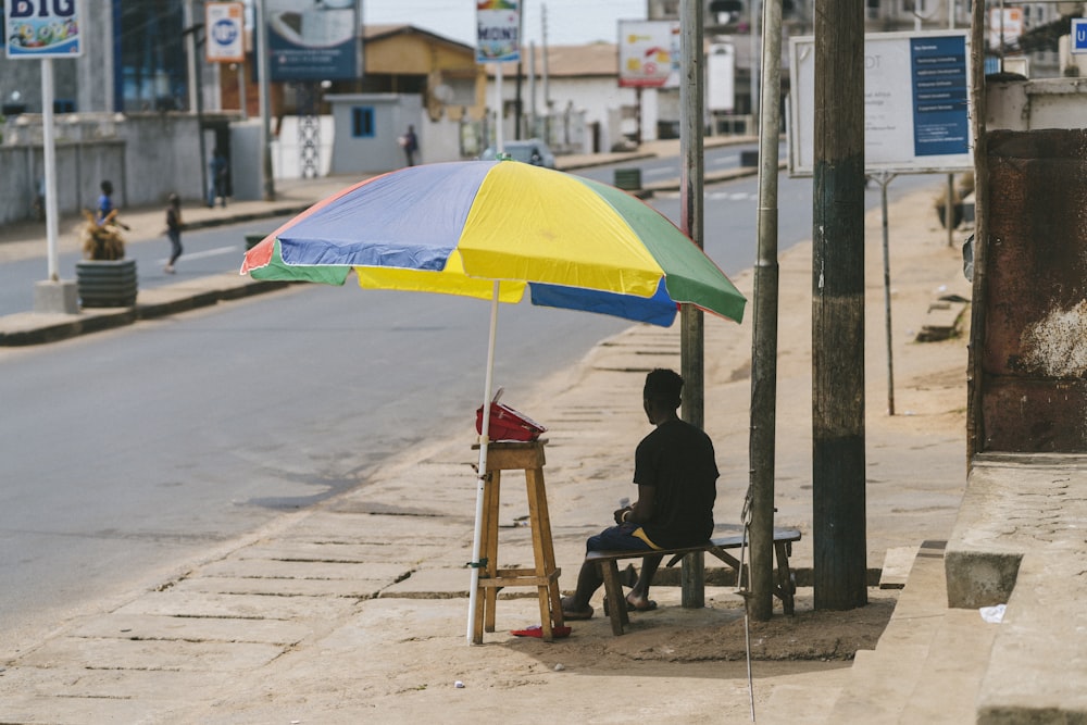 man in black jacket sitting on brown wooden chair under yellow umbrella during daytime