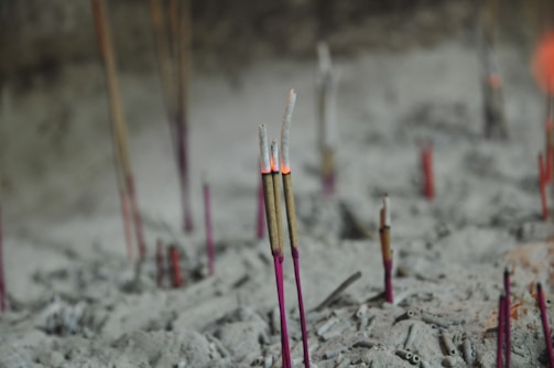 Multiple incense sticks are placed in a sandy or ashen surface with some of them burning and emitting smoke. The setting appears to be a place dedicated to prayer or meditation, as suggested by the presence of numerous incense sticks.