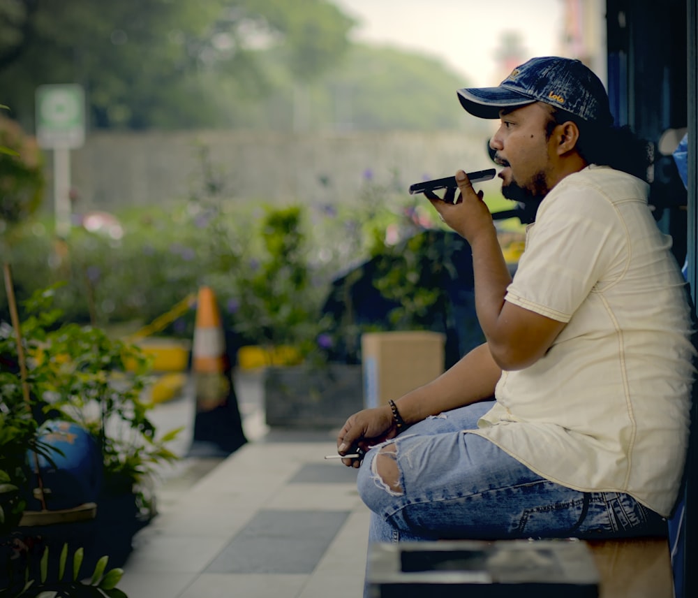 man in white t-shirt and blue denim jeans sitting on concrete bench using black dslr
