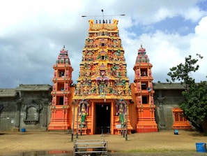 A vibrant and colorful Hindu temple with intricate carvings and statues atop its towers. The central structure features a tall, ornately decorated gopuram with multiple tiers of sculptures depicting deities and mythical figures. The temple is surrounded by a simple stone wall, with greenery and a cloudy sky in the background.