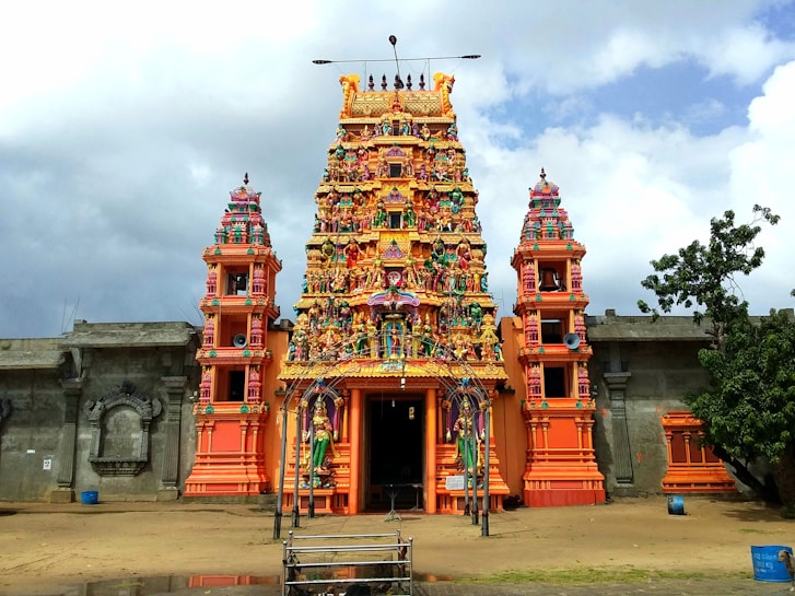 A vibrant and colorful Hindu temple with intricate carvings and statues atop its towers. The central structure features a tall, ornately decorated gopuram with multiple tiers of sculptures depicting deities and mythical figures. The temple is surrounded by a simple stone wall, with greenery and a cloudy sky in the background.