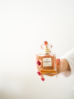 A hand with red manicured nails holding a bottle of Chanel No. 5 Eau de Parfum against a light background. The bottle is clear with a golden cap and contains an amber-colored liquid.