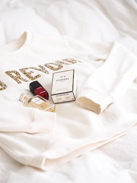 A white sweatshirt with gold glittery text partially visible alongside a red lipstick, a boxed perfume, and a small glass bottle of Chanel No. 5 laying on a soft, white surface.