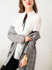 A person with long, wavy hair wears a white blouse with a black tie. They have a grey plaid blazer draped over their shoulders and are wearing a gold watch on their wrist. Their lips are painted red, adding a pop of color to the otherwise neutral tones.