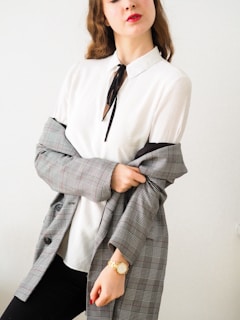 A person with long, wavy hair wears a white blouse with a black tie. They have a grey plaid blazer draped over their shoulders and are wearing a gold watch on their wrist. Their lips are painted red, adding a pop of color to the otherwise neutral tones.