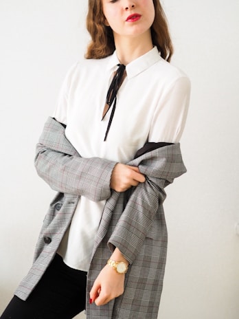 A person with long, wavy hair wears a white blouse with a black tie. They have a grey plaid blazer draped over their shoulders and are wearing a gold watch on their wrist. Their lips are painted red, adding a pop of color to the otherwise neutral tones.