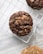 A close-up view of two chocolate cookies with chunks placed on a cooling rack over a newspaper. The cookies have a rustic, homemade appearance with visible chocolate pieces and a textured surface