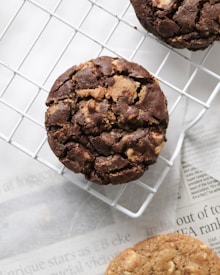 A close-up view of two chocolate cookies with chunks placed on a cooling rack over a newspaper. The cookies have a rustic, homemade appearance with visible chocolate pieces and a textured surface