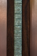 A vertical stack of translucent blue-green glass panes is set within a dark brown wooden frame. The glass has a slightly wavy texture, creating an interesting visual pattern, while the wood provides a contrasting background, highlighting the glass feature.