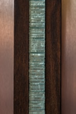 A vertical stack of translucent blue-green glass panes is set within a dark brown wooden frame. The glass has a slightly wavy texture, creating an interesting visual pattern, while the wood provides a contrasting background, highlighting the glass feature.