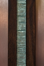 A vertical stack of translucent blue-green glass panes is set within a dark brown wooden frame. The glass has a slightly wavy texture, creating an interesting visual pattern, while the wood provides a contrasting background, highlighting the glass feature.