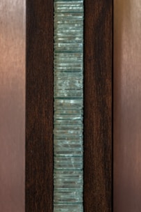 A vertical stack of translucent blue-green glass panes is set within a dark brown wooden frame. The glass has a slightly wavy texture, creating an interesting visual pattern, while the wood provides a contrasting background, highlighting the glass feature.