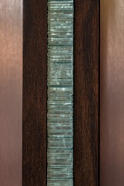 A vertical stack of translucent blue-green glass panes is set within a dark brown wooden frame. The glass has a slightly wavy texture, creating an interesting visual pattern, while the wood provides a contrasting background, highlighting the glass feature.