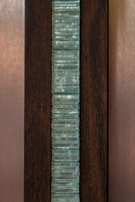 A vertical stack of translucent blue-green glass panes is set within a dark brown wooden frame. The glass has a slightly wavy texture, creating an interesting visual pattern, while the wood provides a contrasting background, highlighting the glass feature.