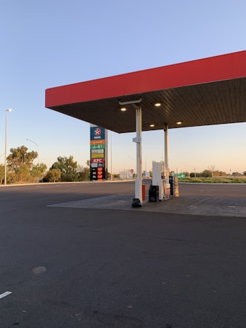 A gas station with a red roof canopy and fuel pumps beneath it is visible. In the background, there is a sign displaying fuel prices and various signs including those for nearby fast-food restaurants. The area is clean and well-lit, with a mostly empty lot and clear skies indicating a quiet and serene atmosphere.