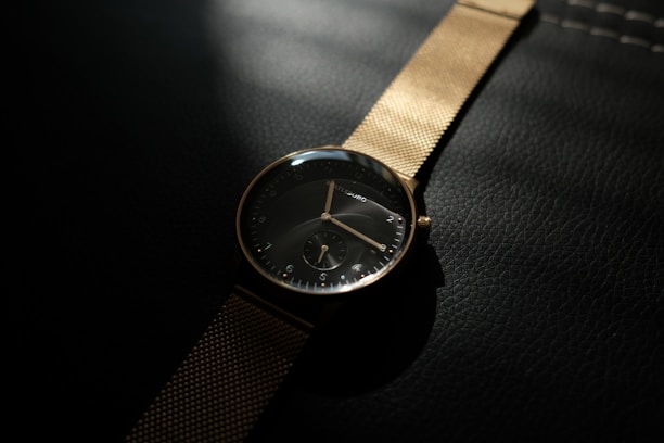 A sophisticated wristwatch with a black face and gold mesh strap lies on a textured dark surface, illuminated by soft lighting. The watch features minimalistic hour markers and a small secondary dial. The composition is elegant, with shadows enhancing the luxurious feel of the watch.